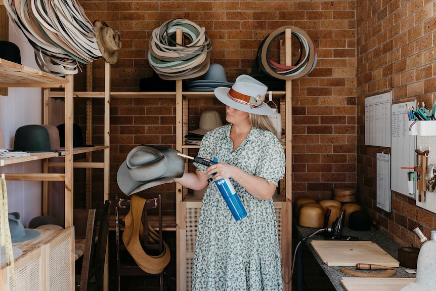 A lady in a hat stands, using a blow torch to style a hat.