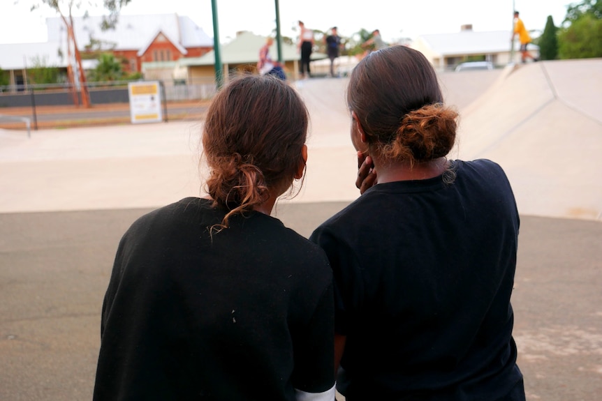 Two girls leaning into each other while they look out over a skate park. 