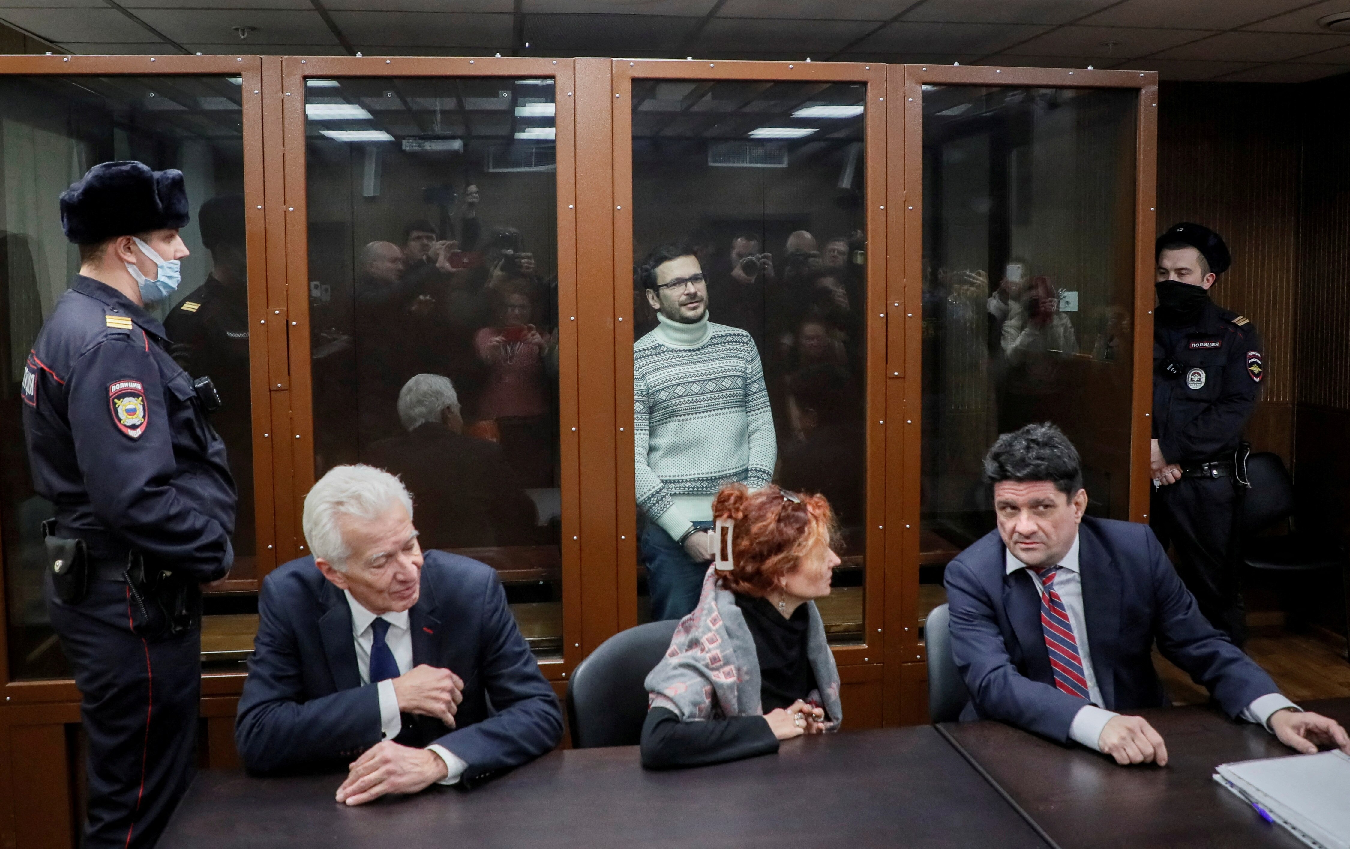 Ilya Yashin stands inside glass cage inside court as lawyers sit in front of glass and guards stand either side.