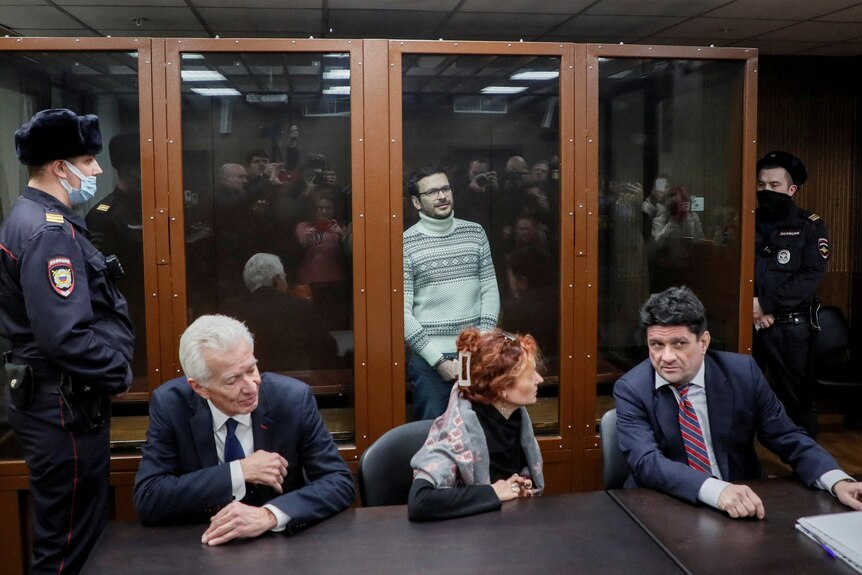 Ilya Yashin은 법원 내부의 유리 우리 안에 서 있고 변호사는 유리 앞에 앉아 있고 경비원은 양쪽에 서 있습니다.