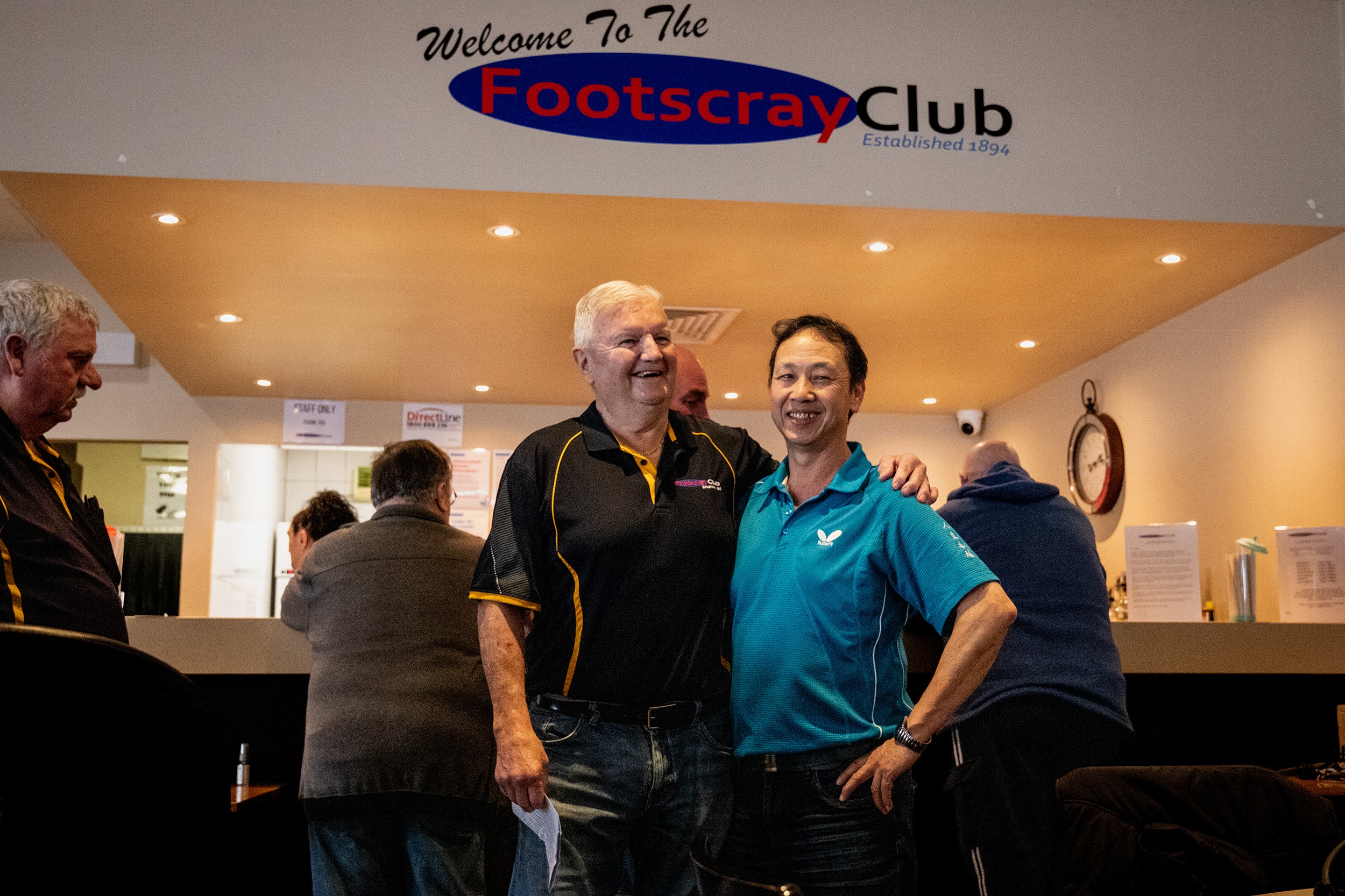 Two men, one wearing a black polo and the other a blue polo, stand smiling with arms around one-another in front of a bar