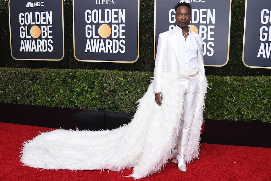 Actor Billy Porter wearing a white tuxedo with a long, feathered train the trials behind him.