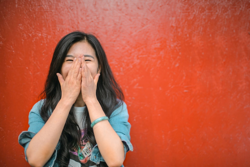 A woman of Asian heritage stands against a red wall, holding her hands to her face, laughing