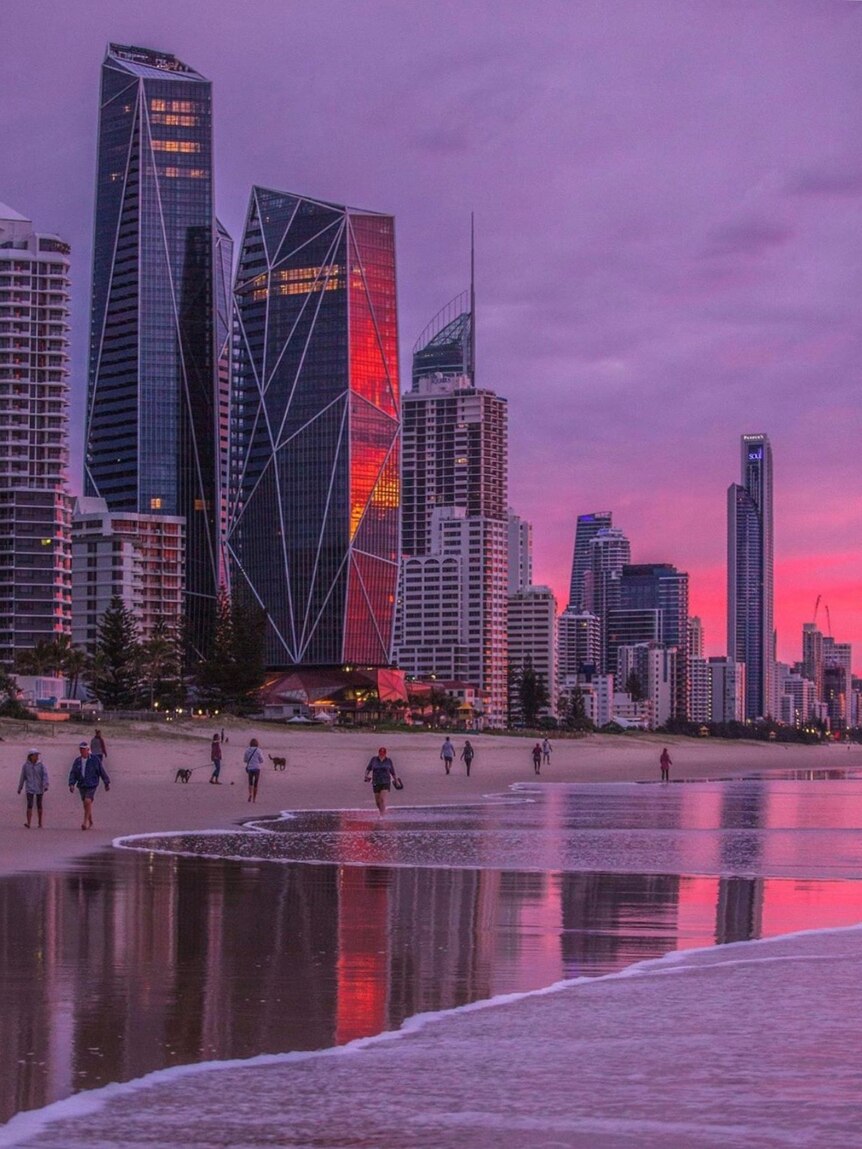 People walk on the beach under the Gold Coast skyline with a pink and purple sunrise.