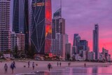 People walk on the beach under the Gold Coast skyline with a pink and purple sunset.