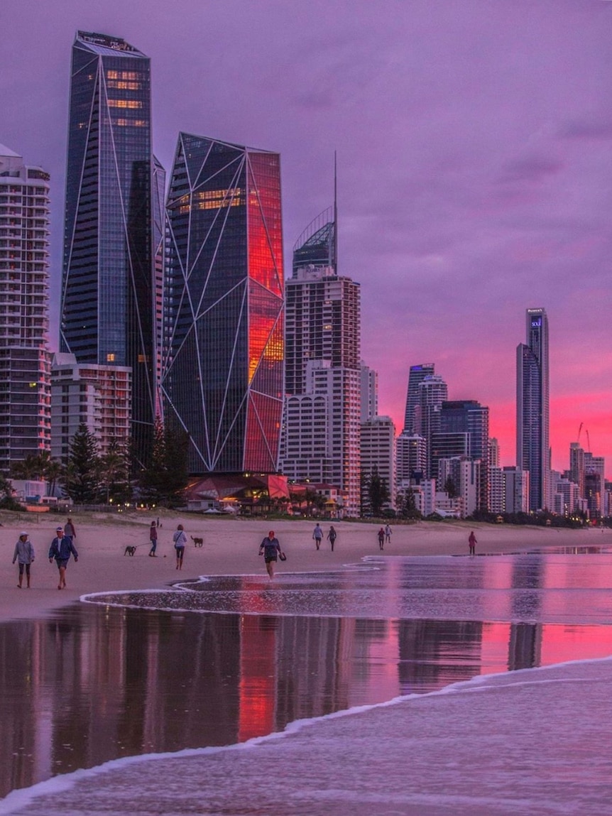 People walk on the beach under the Gold Coast skyline with a pink and purple sunset.