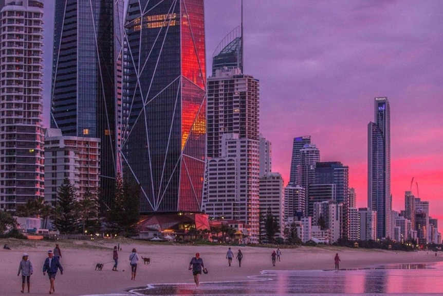 People walk on the beach under the Gold Coast skyline with a pink and purple sunrise.