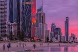 People walk on the beach under the Gold Coast skyline with a pink and purple sunset.