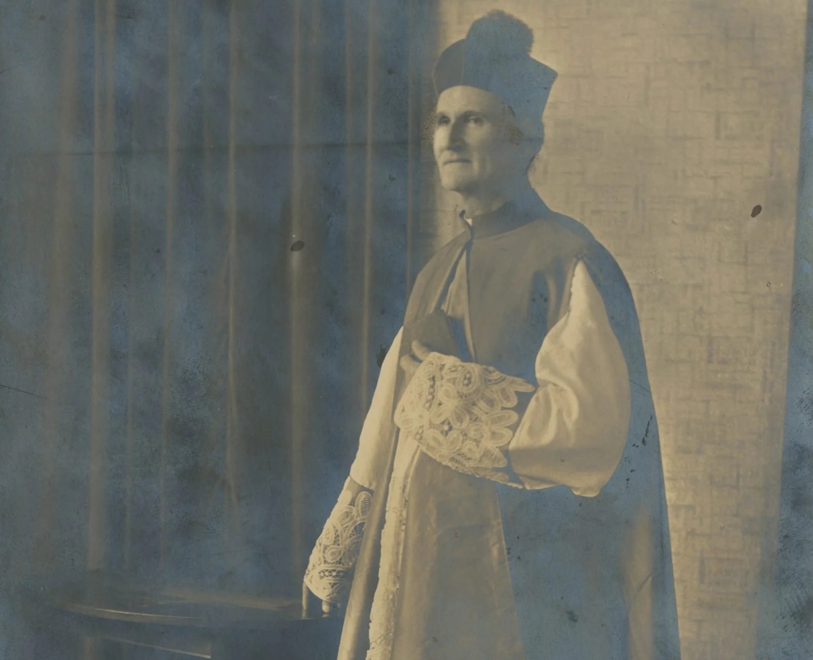 John Hawes wears a long-sleeved priest outfit, holding a small book and looks off to the distance.