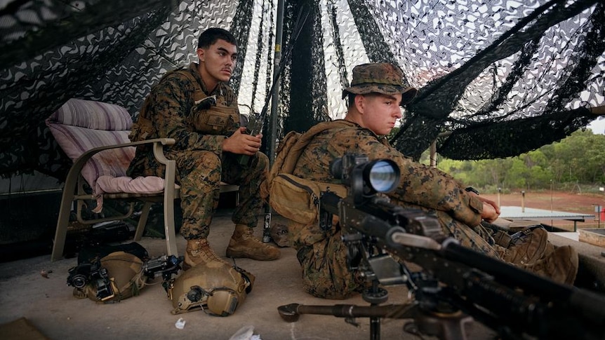 Two marines in army gear sitting in a tent, a rifle next to them