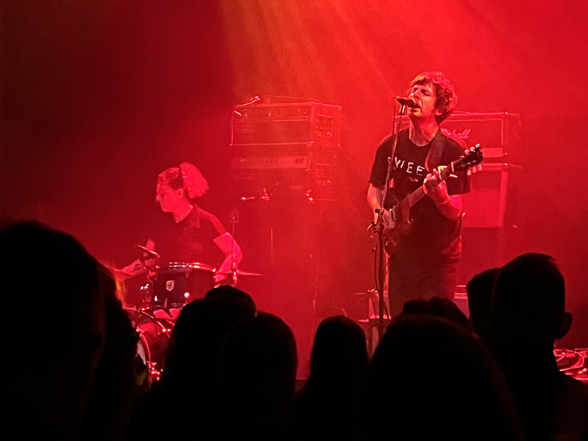 Man and women play guitar and drums on stage with red backdrop