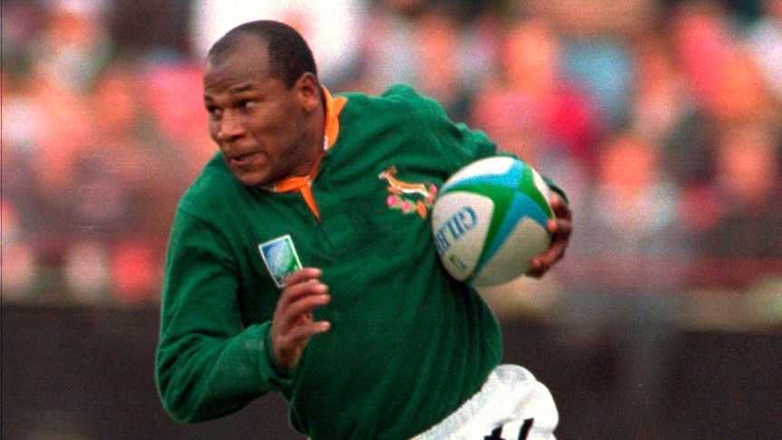 Chester Williams carries the ball under his left arm and runs, wearing a green springbok jersey