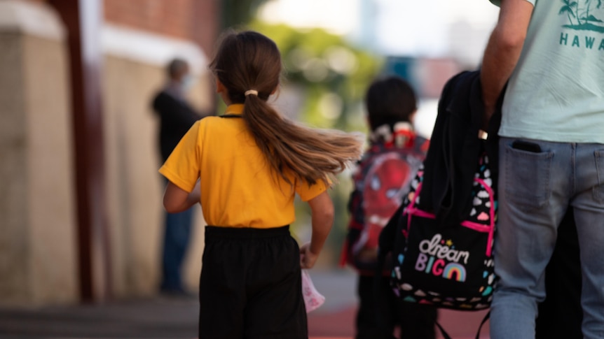 Girl in yellow top walks while her dad carries her schoolbag.