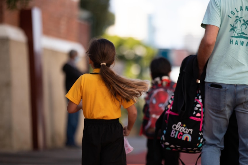 Girl in yellow top walks while her dad carries her schoolbag.