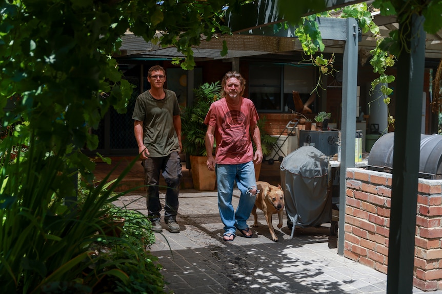 Two men walk with a dog through a backyard patio surrounded by ferns