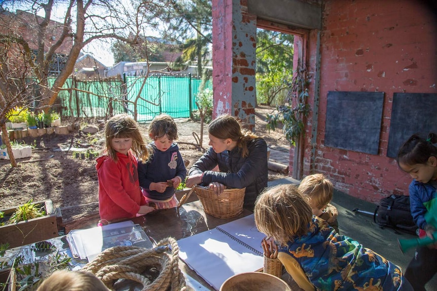 Children and an adult gather around a table covered in baskets, rope, a sketchbook, and plant samples.
