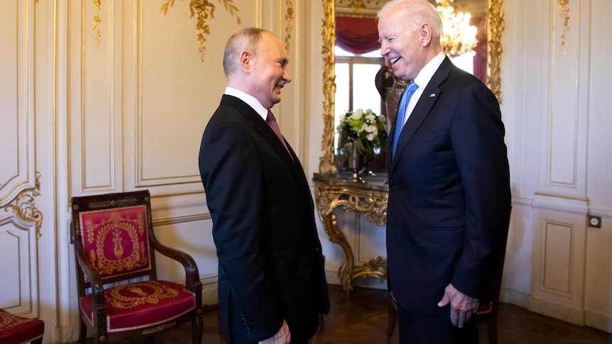 Joe Biden and Vladimir Putin in dark suits smile at one another while standing in a room.