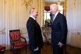 Joe Biden and Vladimir Putin in dark suits smile at one another while standing in a room.