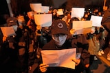 Protesters holding up blank pieces of paper in a crowd. Many are wearing caps and masks. 
