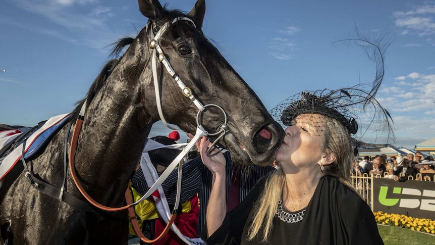 A woman wearing a feathered hat kisses a horse's face.