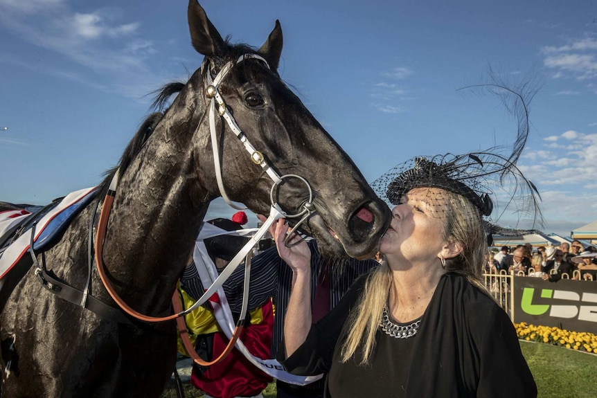 A woman wearing a feathered hat kisses a horse's face.