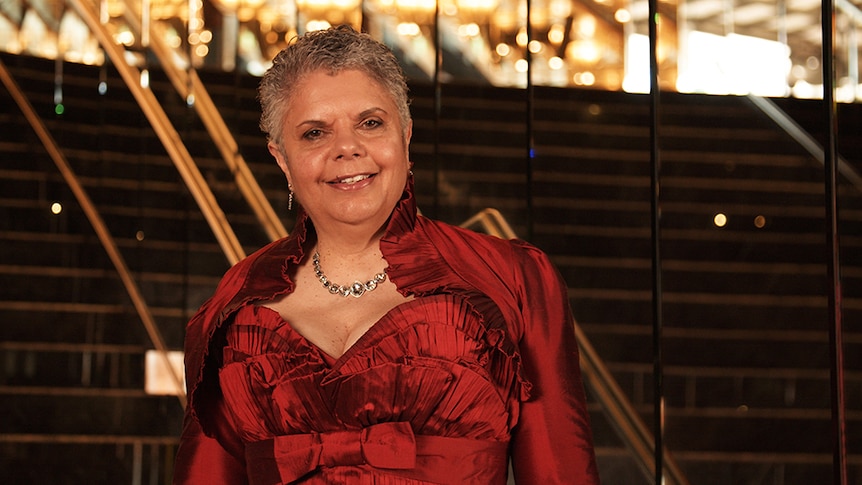 Yorta Yorta composer and soprano Deborah Cheetham stands on stairs surrounded by mirrors in a red evening gown.