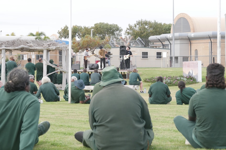 Inmates dressed in green sit and watch a band perform outside in a grassed area.