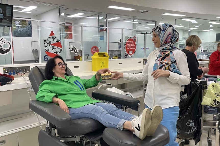 A woman standing next to another woman laying in a chair about to donate blood, handing her a chocolate.
