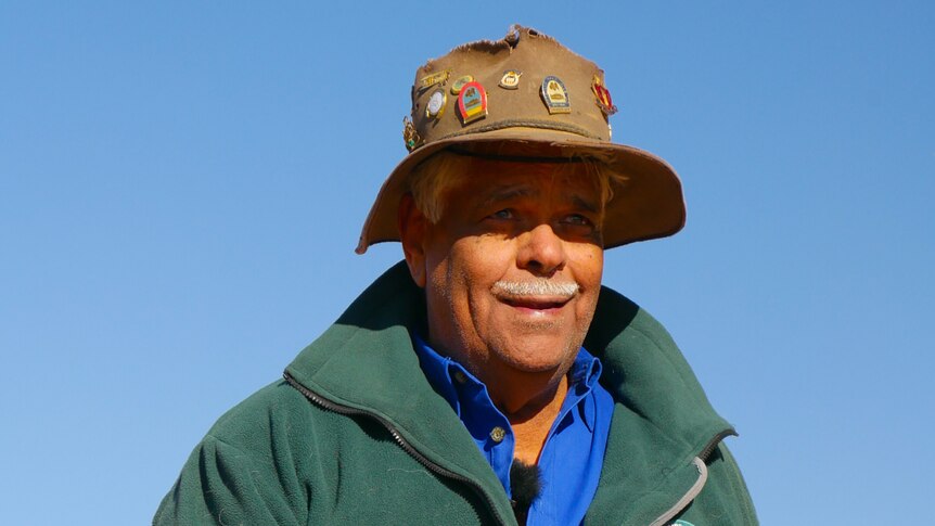 An Indigenous man wearing an Akubra-style hat with badges, and a Parks and Wildlife jacket, holds a rock in the desert.