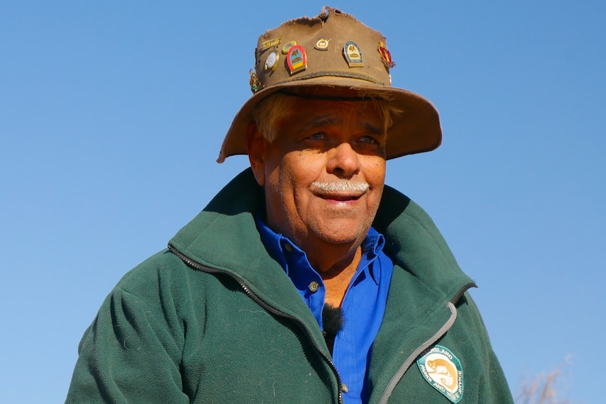 An Indigenous man wearing an Akubra-style hat with badges, and a Parks and Wildlife jacket, holds a rock in the desert.