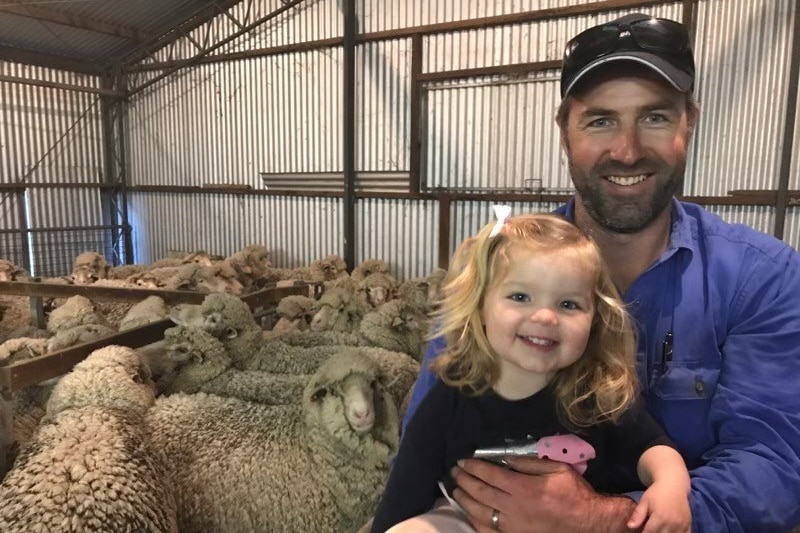 A man in a blue shirt smiles holding a young girl next to a pen full of sheep