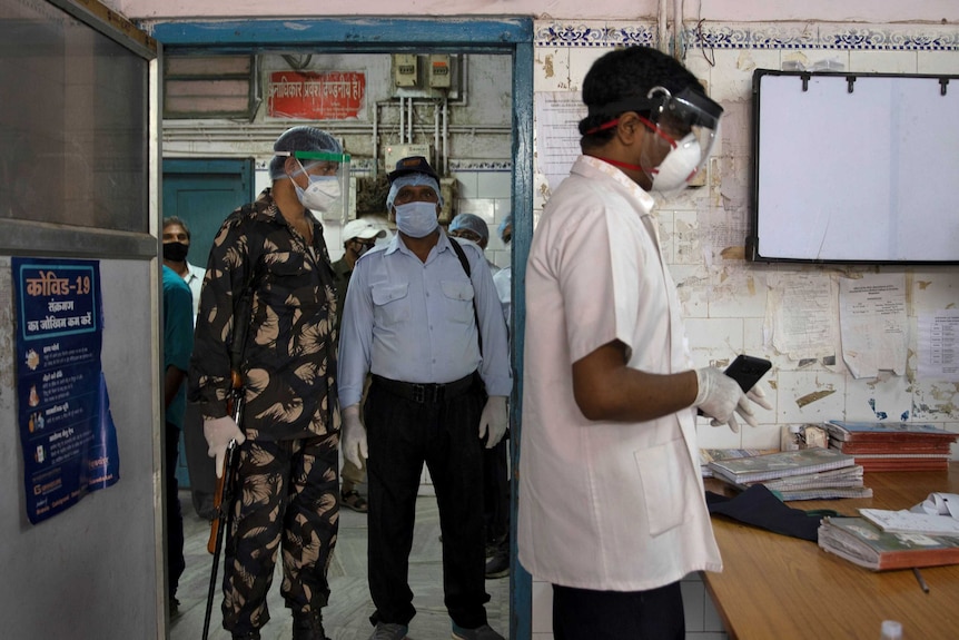 A doctor wearing a white shirt and a mask and face shield stands in a room while an armed man stand guard holding a gun.