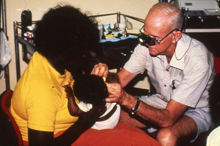 An Aboriginal woman holding a baby in her lap as a white man with special glasses looks in the baby's ear.