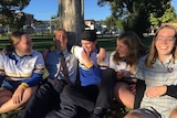 A boy sitting on ground, wearing a protective skull cap, surrounded by three girls and a boy, all in school uniform