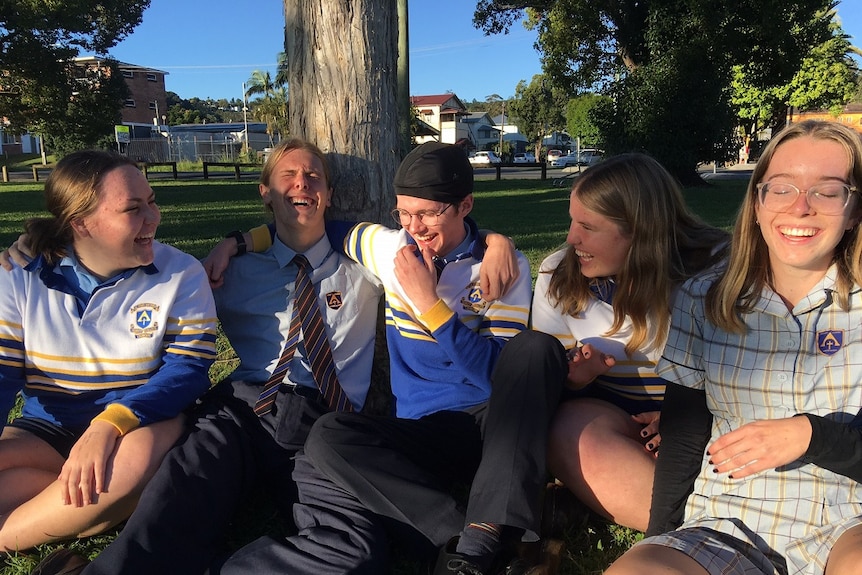 A boy sitting on ground, wearing a protective skull cap, surrounded by three girls and a boy, all in school uniform