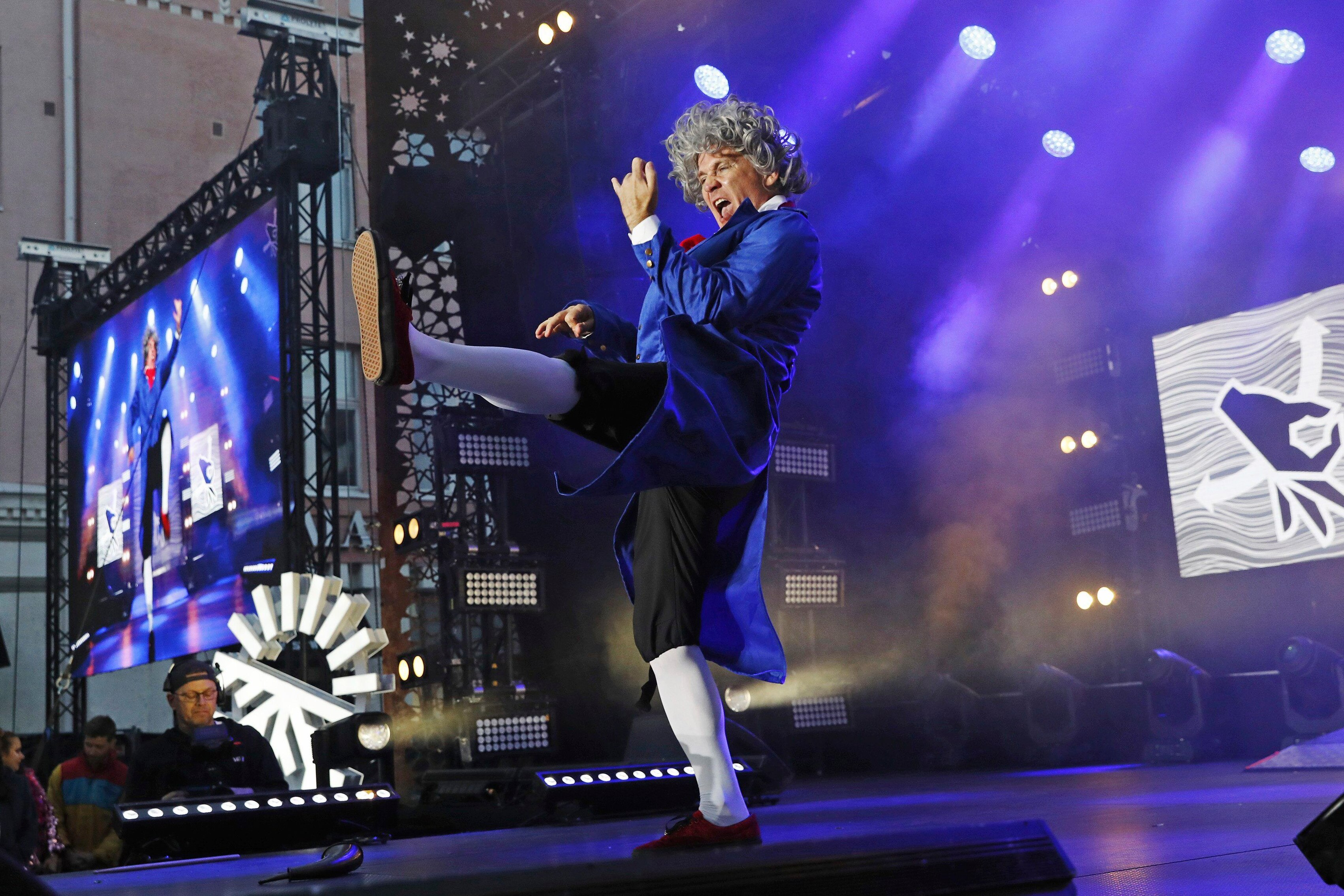 A man in an old fashioned blue suit and grey wig performing air guitar on bright blue stage.
