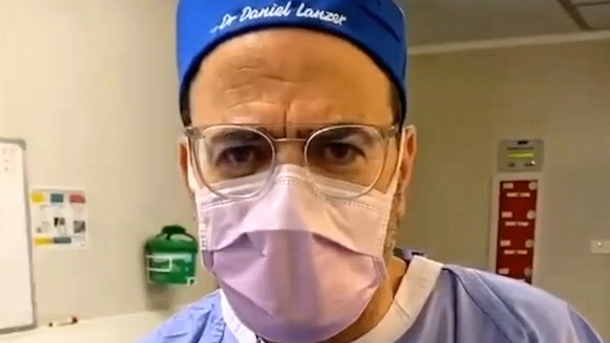 Dr Lanzer looks at the camera. He is dressed for surgery, wearing a mask, gown and a hat with his name embroidered on it.