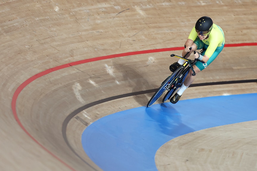 A cyclist in a green and gold lycra cycling uniform and a helmet rides around one of the corners of a velodrome.