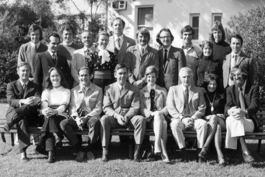 A black and white group photo of 19 people, men and women, dressed in office attire of the 1970s.