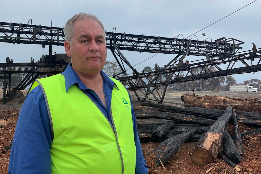 a picture of Malcolm McComb in a high vis jacket standing near a burnt out crane and logs