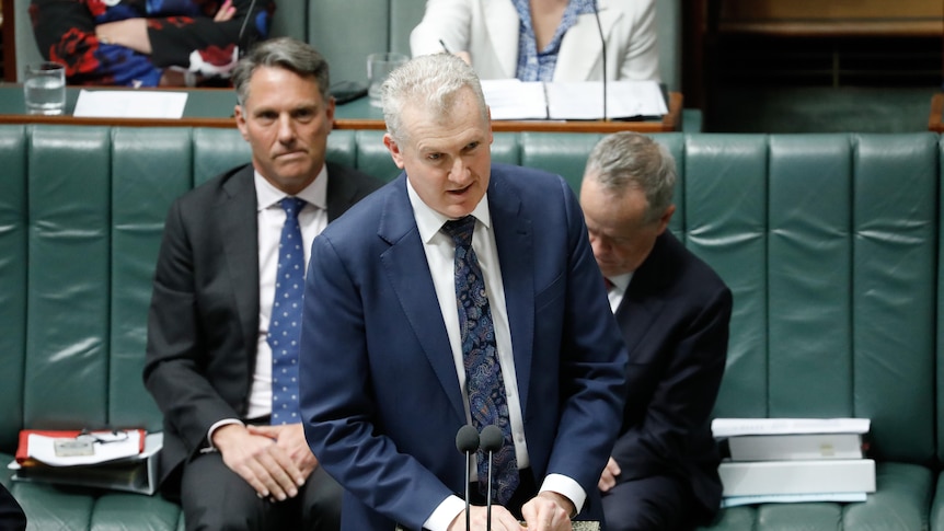 Tony Burke, a man wearing a blue suit and print tie, speaks into a microphone in front of two colleagues