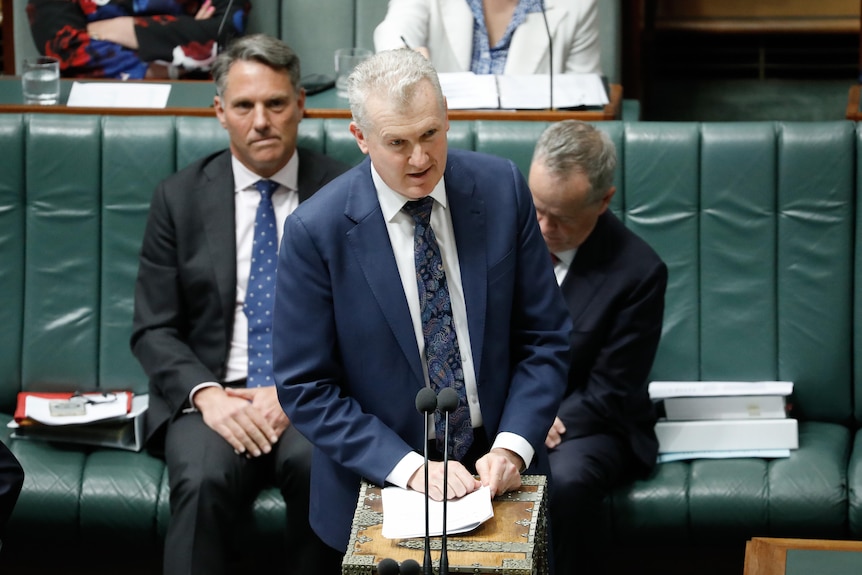 Tony Burke, a man wearing a blue suit and print tie, speaks into a microphone in front of two colleagues