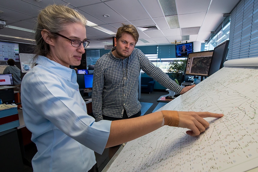 A woman points out weather patterns on a map, showing another man. 