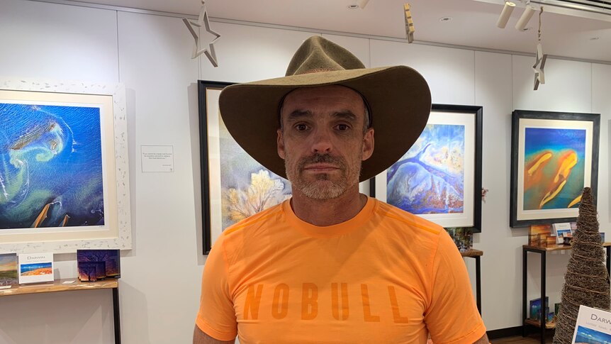 A man in a large brown hat stands in an art gallery. 