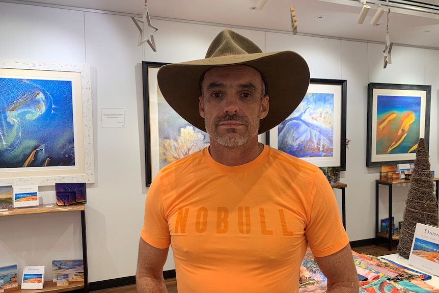 A man in a large brown hat stands in an art gallery. 