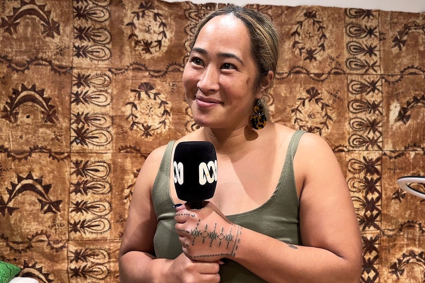 Woman with hair tied back in ponytail holds ABC microphone with tualima, Samoan hand tatau freshly inked. Wears green singlet.