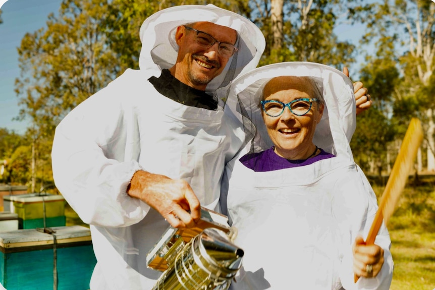 A man and a woman wearing beekeeper suits hold a bee smoker.