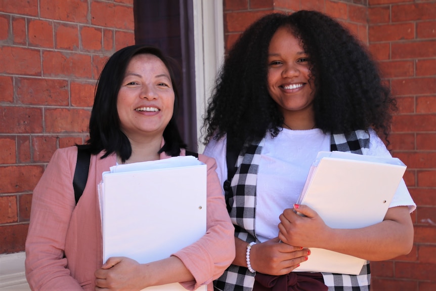 Two women with dark hair hold folders in front of a red brick building.
