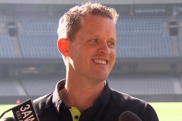 A man with brown hair smiles as he answers questions from reporters in front of an empty stadium.