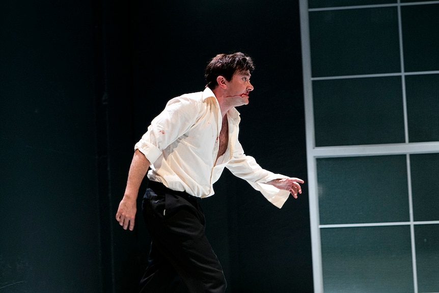 A dishevelled man in half unbuttoned beige shirt with blood running down face from nose walks across theatre stage.